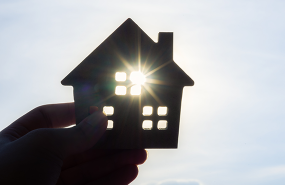 A hand holds the wooden cutout of a house up to the sun, and the light streams through the cutout windows of the house.