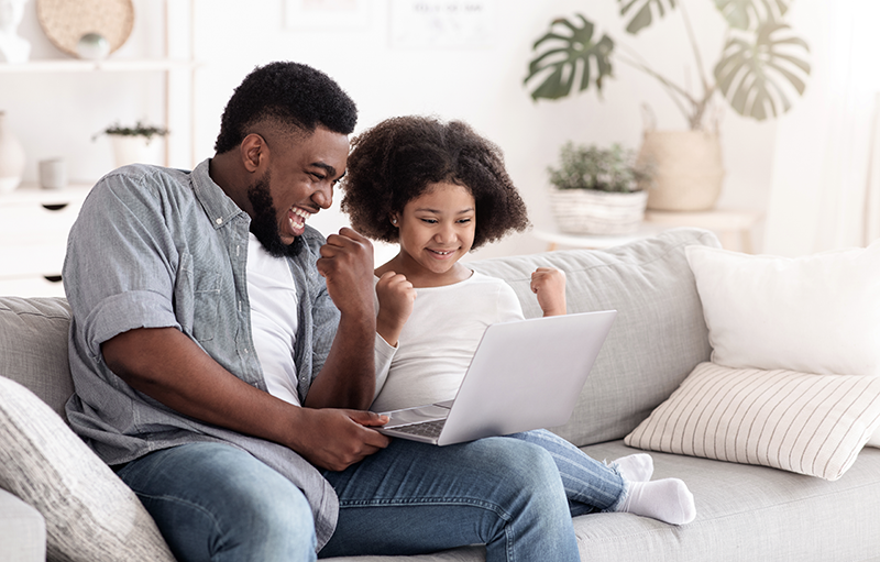 A father and his daughter sit together on the couch, visibly excited about something they see on the laptop they're sharing
